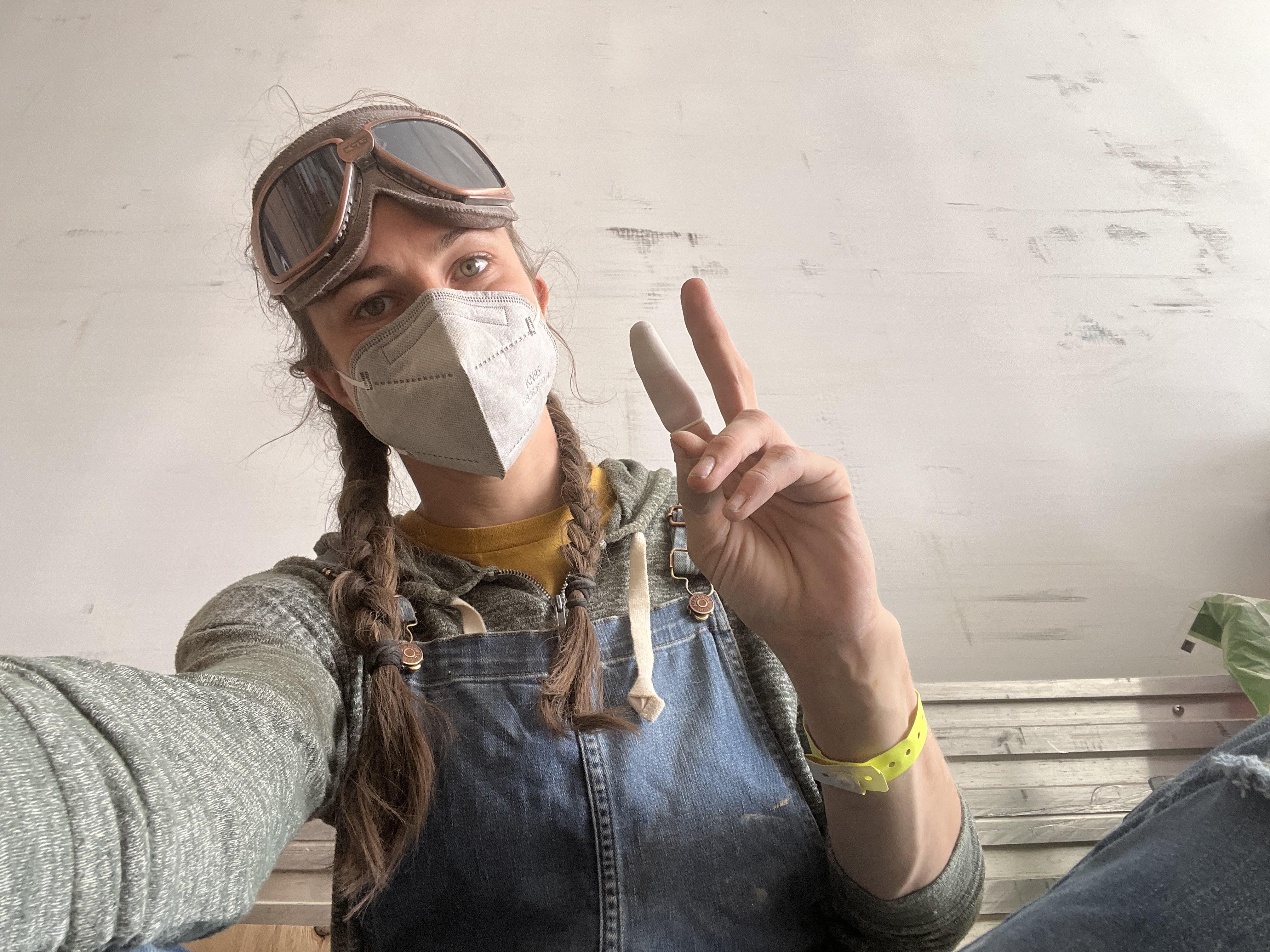 me with a mask and goggles on, hair braided, putting up peace sign with hands